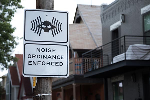 Noise Ordinance Enforced sign with a symbol of person covering ears, posted with a neighborhood houses in the blurry background